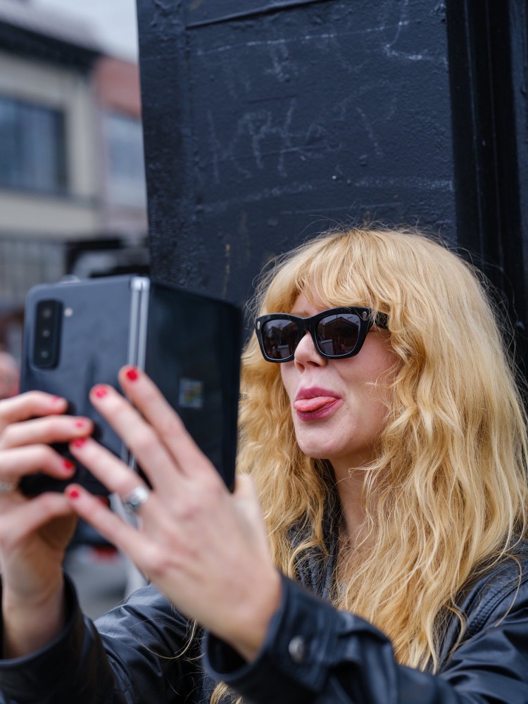A woman in dark sunglasses holds a phone up to take a selfie, tongue out, shot with flash