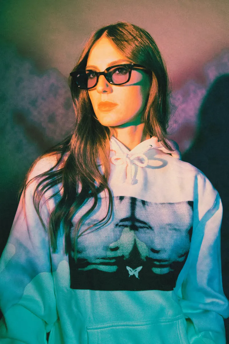 A woman in tinted glasses and a white CLSICS hoodie, lit in teal and violet