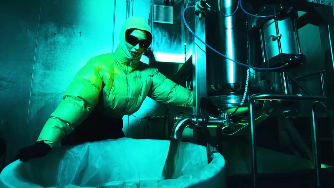A woman in a green puffer jacket and dark glasses works a stainless steel tank in a teal-lit rosin lab