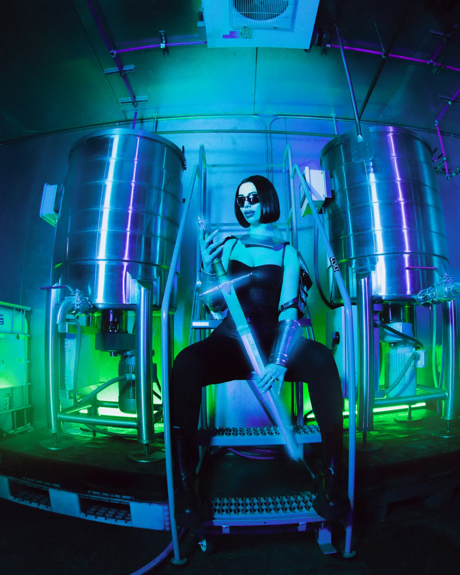 A woman in dark glasses and a black bodysuit sits between two steel tanks in a blue and green lit rosin lab