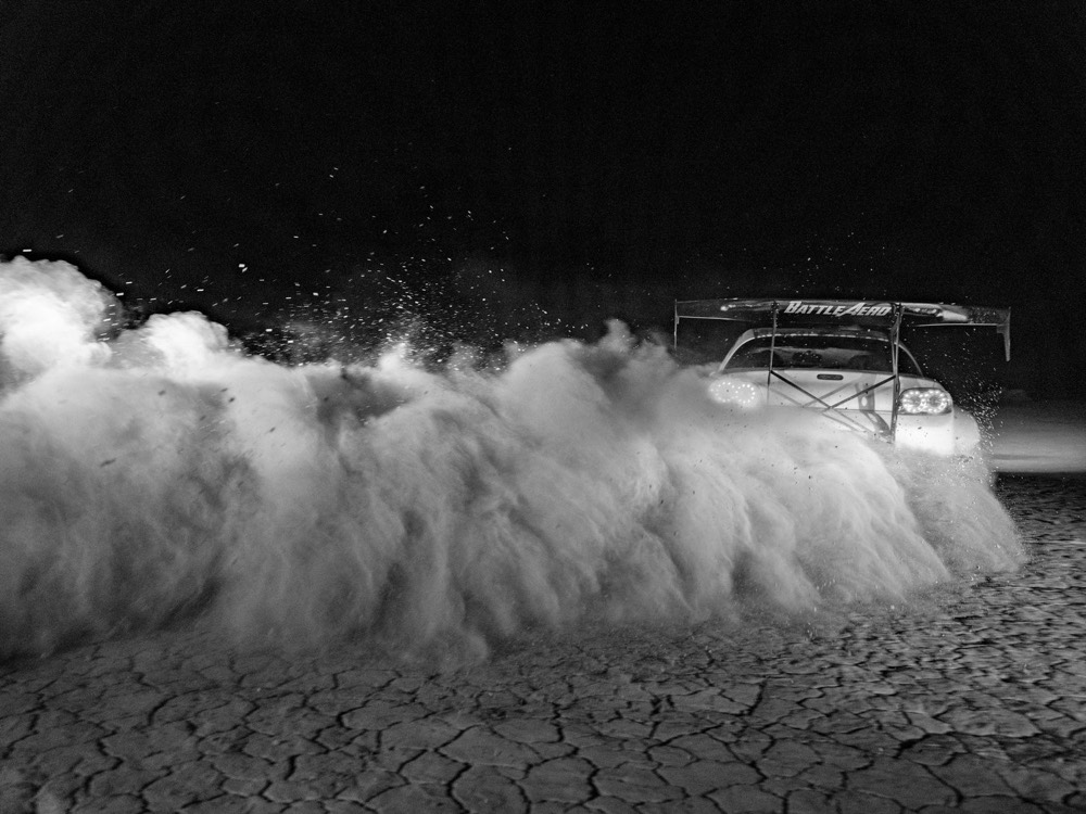 Black and white photograph of a car throwing a wall of dust across a dry lake bed