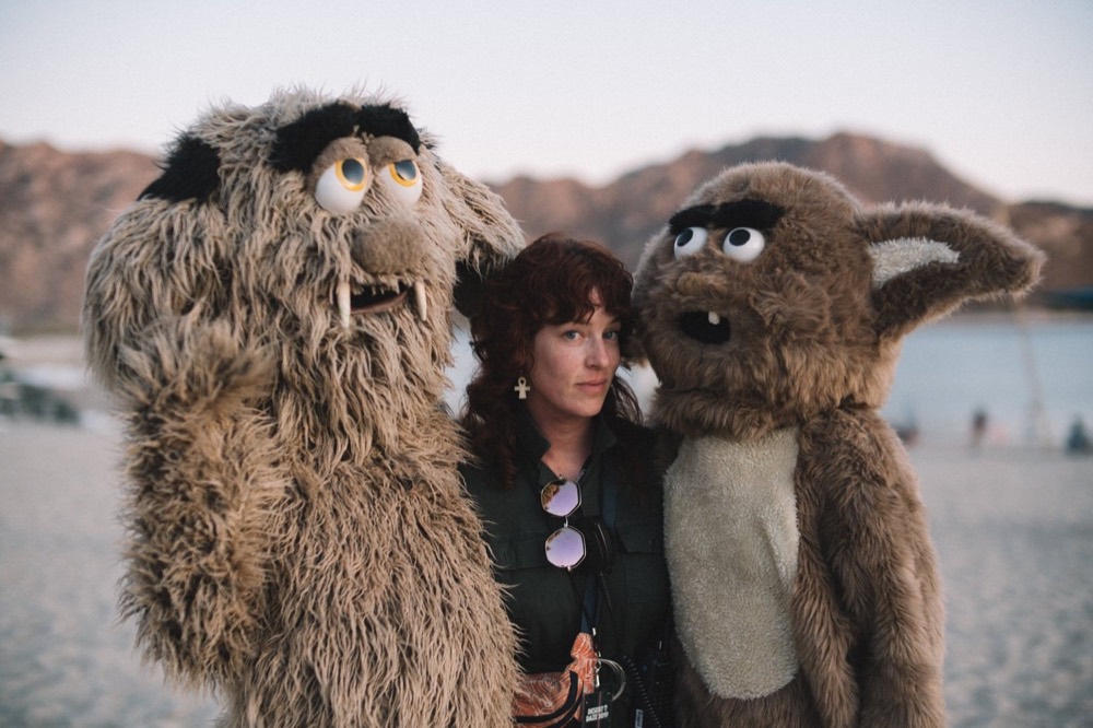 A woman on a beach flanked by two people in large shaggy creature costumes