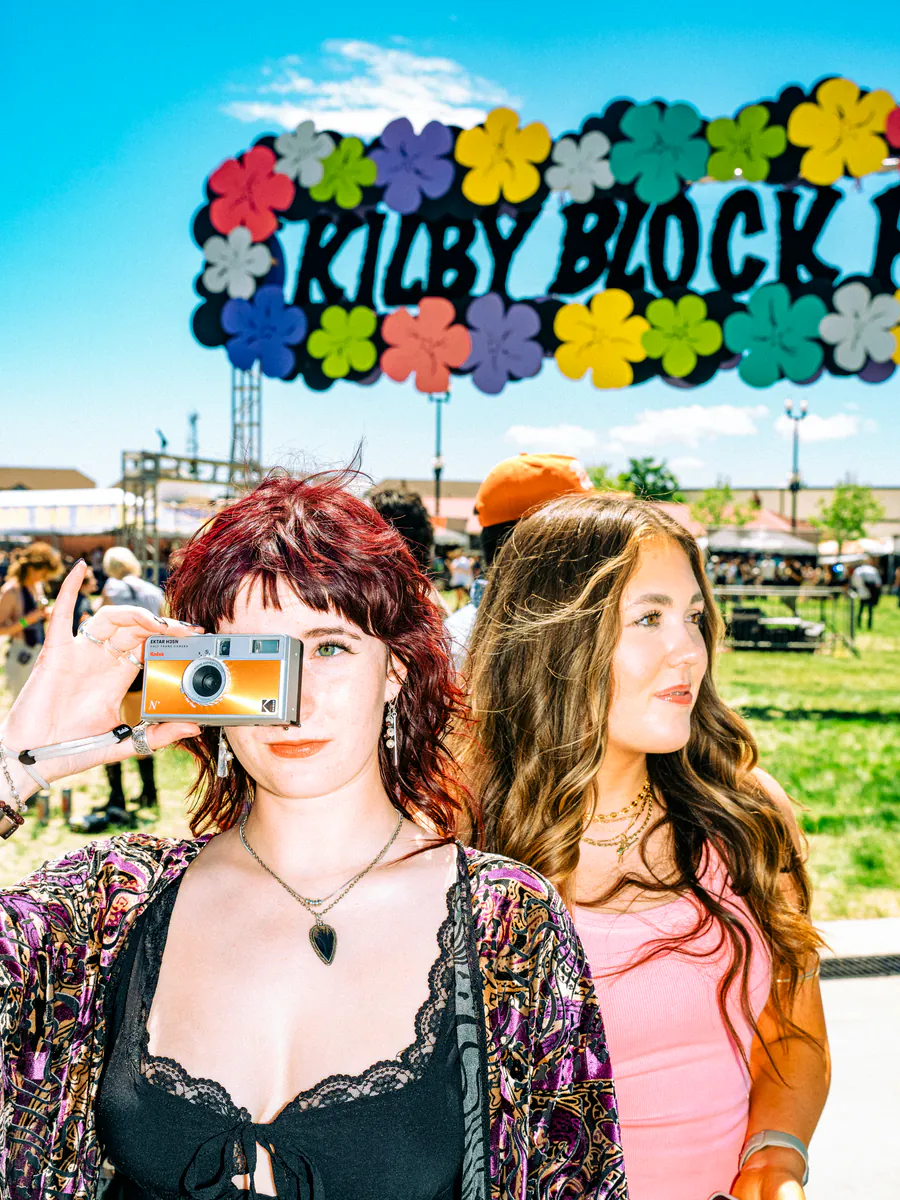 A red-haired festival-goer takes a photo on a film camera beside a friend, under the flower-covered Kilby Block Party sign