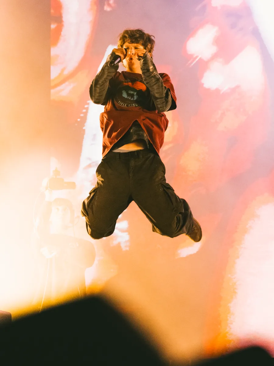 A singer leaps high above the stage, knees tucked, against a glowing orange screen and stage haze