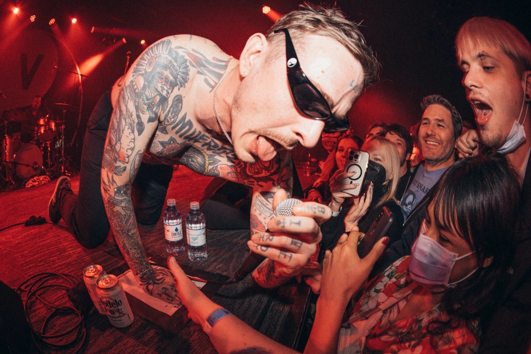 A tattooed singer leans down into a packed crowd, phones raised, lit red