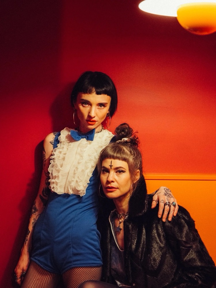 Two women posed against a red and orange wall, one in a blue dress, shot with hard flash