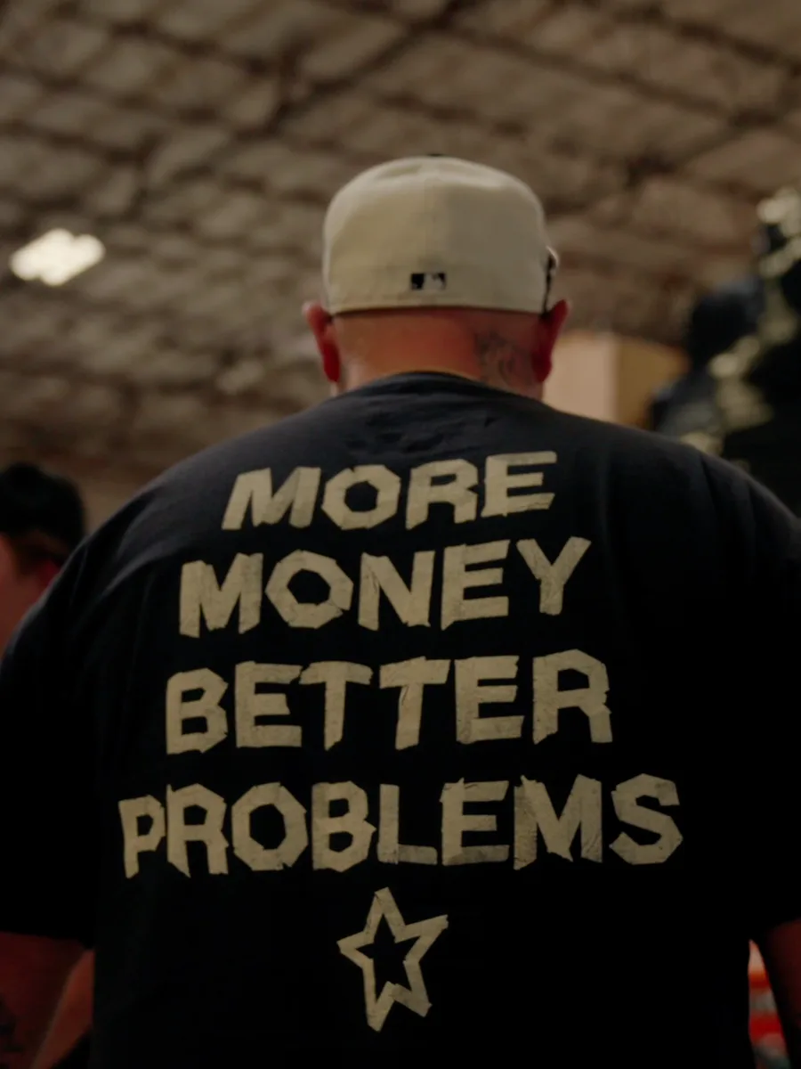A man in a cap, seen from behind in a warehouse, wearing a black YNGSTAR tee that reads "More Money Better Problems"