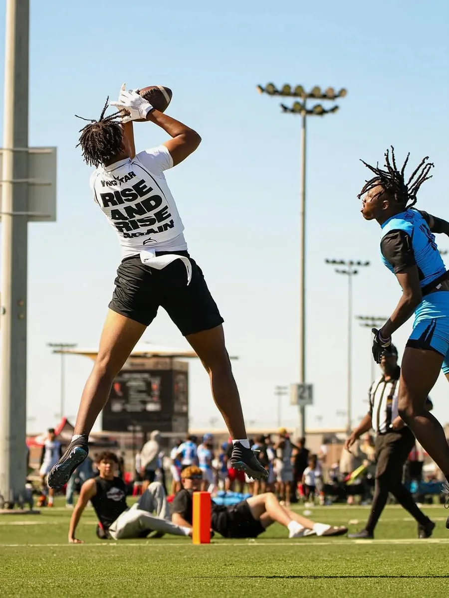 A player in a YNGSTAR "Rise and Rise Again" tee leaps to catch a football as a defender closes in on a sunny tournament field