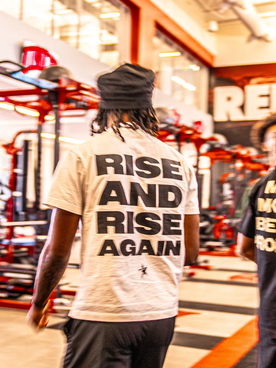 A man walking through a red-racked gym, seen from behind in a white YNGSTAR tee that reads "Rise and Rise Again"