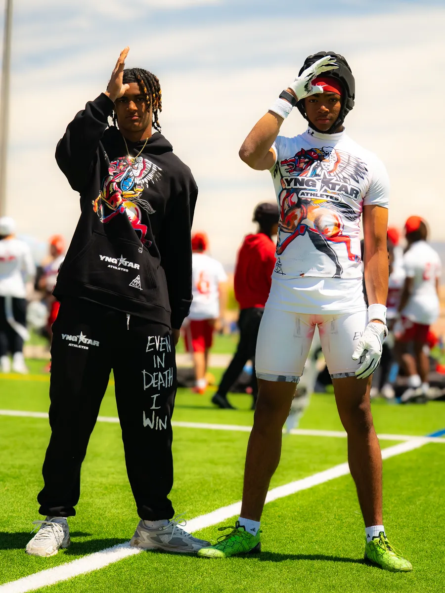 Two young athletes posing on the turf, one in black YNGSTAR sweats reading "Even in Death I Win", the other in a white YNGSTAR kit and helmet